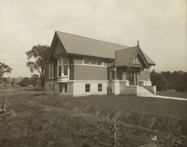 Exterior view of the Kaukauna Public Library. The library opened in 1905 and was funded by Andrew Carnegie with a gift of $12,000. Visible above the main entrance are the words: "Public Library." The reverse of the cardboard mount reads: "Kaukauna Wis., 1905." Brick building with decorative wood trim along the roof line. In the background are trees, and the lawn area in front of the library is elevated above the surrounding land.