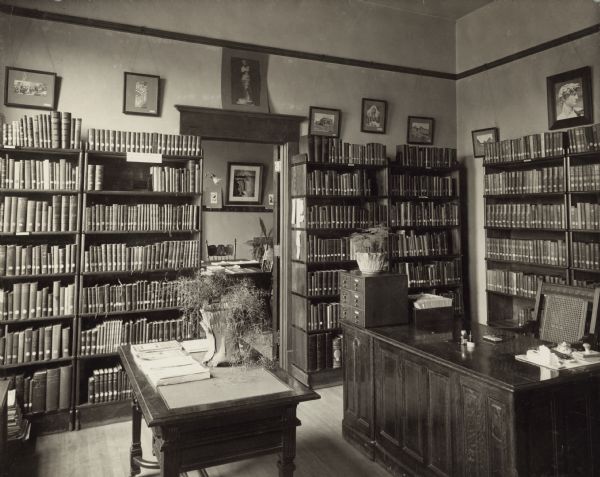 Interior view of the Kaukauna Public Library. The library opened in 1905 and was funded by Andrew Carnegie with a gift of $12,000. In the room is a librarian's desk, a small six-drawer card catalog, and bookshelves along the walls. An open doorway leads into another room with a reading table visible.