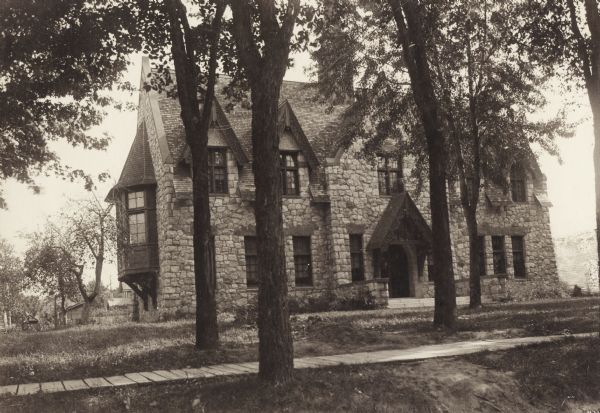 Exterior view of the Fargo Public Library in Lake Mills. Reverse of cardboard backing reads: "1902, $8000 - Cost, G.B. Ferry, Milwaukee, arch." The library is a stone building with intricate detailing on the gables and over the door. A wood plank sidewalk is in the foreground.
