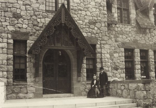 Exterior view of the Fargo Public Library in Lake Mills. Visible above the main entrance are the words: "Public Library." Reverse of cardboard backing reads: "1902, Cost $8000 - G.B. Ferry, Milwaukee, arch." In front of the entrance, which has an intricately carved roof, a man is standing near a seated woman.