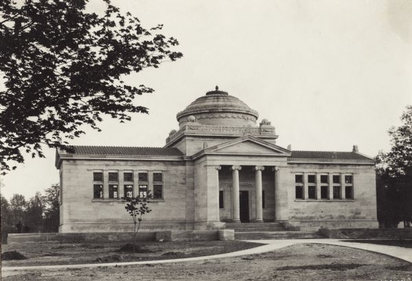 Exterior view of the Gilbert M. Simmons Public Library. The library opened in 1900 and was funded with a gift of $150,000 from G.M. Simmons. The building is of cut stone, columns are in front of the porch entrance, and a dome is in the center of the roof. The grounds are newly landscaped and sidewalks lead up to the entrance.