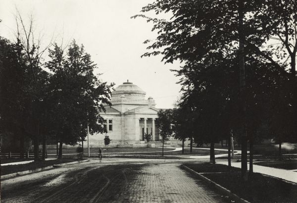 Exterior view from tree-lined cobblestone street of the Gilbert M. Simmons Public Library. The library opened in 1900 and was funded with a gift of $150,000 from G.M. Simmons. Two bicyclists are on the street on the left.