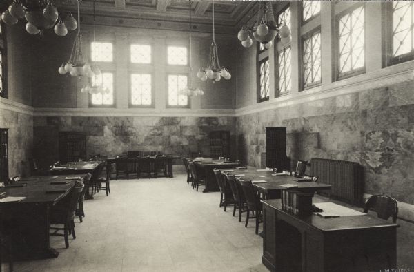 Interior view of the Gilbert M. Simmons Public Library. The library opened in 1900 and was funded with a gift of $150,000 from G.M. Simmons. On the right is a librarian's desk, and reading tables and chairs are along the edges of the room. A few freestanding bookshelves are distributed along the marble walls of the room. Large chandeliers hang from the high ceiling, and large windows are on all three sides of the room.