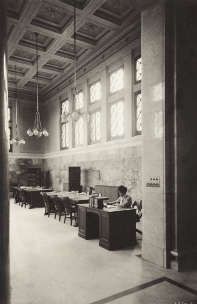 Interior view of the Gilbert M. Simmons Public Library. The library opened in 1900 and was funded with a gift of $150,000 from G.M. Simmons. Through a large opening a woman (librarian?) can be seen seated at a librarian's desk, in a room with reading tables and bookshelves. There is a high, ornate coffered ceiling, with chandeliers suspended from it, large windows, and marble walls.