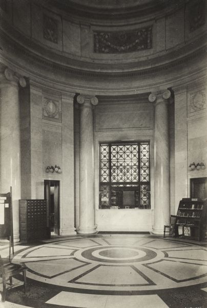 Interior view of the rotunda of the Gilbert M. Simmons Public Library. In the center background is a window with a sign that reads: "Renting Collection." A sign that reads: "New Books" is visible. Lining the walls of the rotunda are a card catalog, a bookshelf and chairs.
