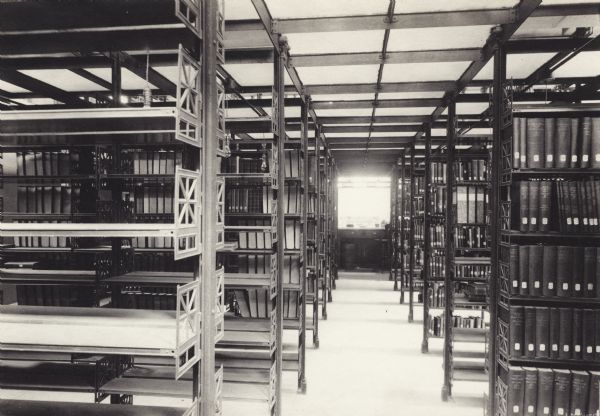 Interior view of the Gilbert M. Simmons Public Library. Reverse of cardboard mounting reads: "1899, cost $150,000, D. H. Burnham, Chicago, Arch." A second note, in blue, reads: "Return to Wisconsin Library School. Stationary bookshelves on either side of an aisle leading to a table and chair in a room with a low ceiling.