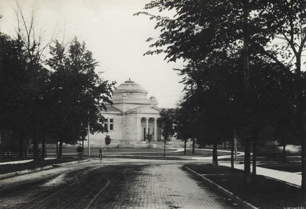 Exterior view from tree-lined cobblestone street of the Gilbert M. Simmons Public Library. The library opened in 1900 and was funded with a gift of $150,000 from G. M. Simmons. Two bicyclists are on the street on the left.