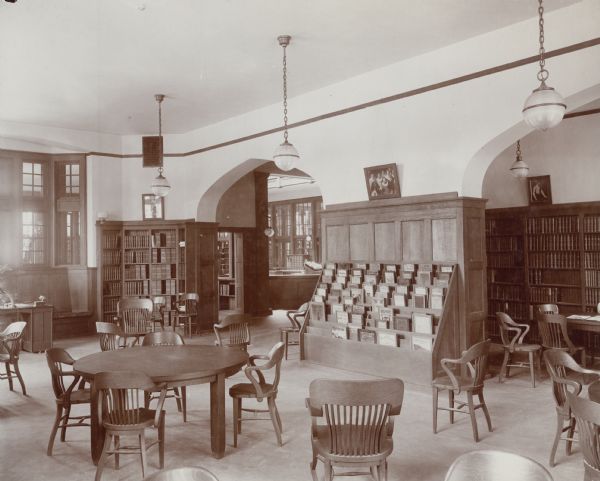 Interior view of the Madison Public Library. The library opened in 1905 and was funded by a $75,000 gift from Andrew Carnegie. Reverse of cardboard backing reads: "Madison (Wisconsin) Free Library reading room, taken from front end. Shows part of the study alcove, corner of Reference Room, charging desk and offices back of catalog case. Bound periodicals are shelved in Reading Room, alphabet starting in corner of alcove shown, going straight around the room and ending in corner between entrance and window seat, where also are shelved boxes of current periodicals."