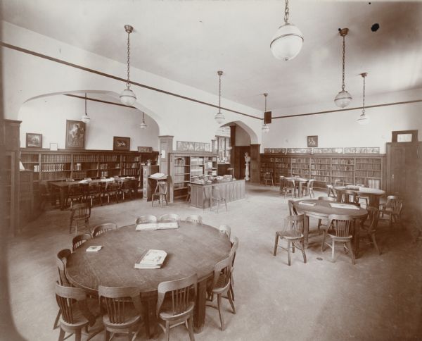 Interior view of the Madison Public Library. The library opened in 1905 and was funded by a $75,000 gift from Andrew Carnegie. Reverse of cardboard mounting reads: "Madison (Wis.) Free Library Children's Room taken from front end. Door at the right opens into cloak closet, alcove used as classroom for teachers; has children's reference books for grade work in schools, pedagogical books for teachers, card catalogue, dictionary stand, and picture case. The frieze across end of room is continued all around the room. It is made of the Walter Crane books, each story being complete in one frame. Fiction is shelved at the rear of the room; classed books along the side wall (not shown); and picture books and books for the very little people in the corner case in front, between the catalog case and the window seat (not shown)."