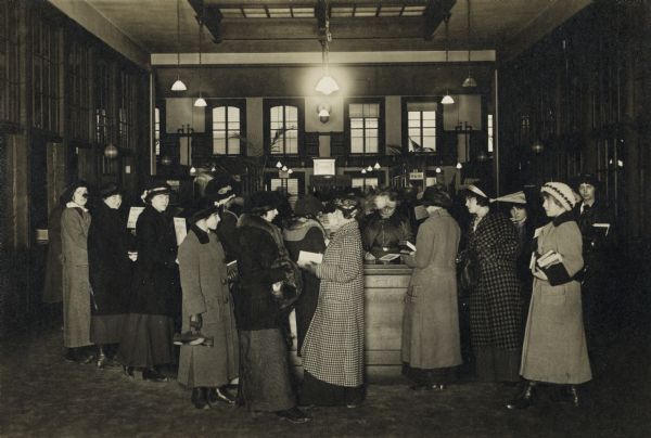 Interior view of the Madison Public Library, circulation desk. Cardboard backing reads: "1913, Main Loan Desk." A large group of women in winter coats and hats are standing around the desk, holding books. A librarian is at the loan desk.