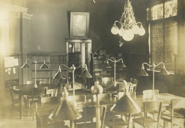 Interior view of the Stephenson Public Library. The library opened in 1903 funded with a gift from Isaac Stephenson of $30,000. The reading tables have large lamps installed on them, bookshelves and a display rack are on the back wall. A large chandelier hangs from the ceiling, and a doorway opens into another room. A framed image of Michelangelo's David hangs above the door.