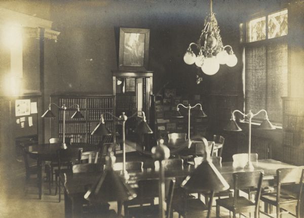 Interior view of the Stephenson Public Library. The library opened in 1903 funded with a gift from Isaac Stephenson of $30,000. Reverse of mounting cardboard reads: "Stephenson Public Library - Marinette Wis. Adult reading room." The reading tables have large lamps installed on them, bookshelves and a display rack are on the back wall. A large chandelier hangs from the ceiling, and a doorway opens into another room, and a framed image of Michelangelo's David hangs above the door.