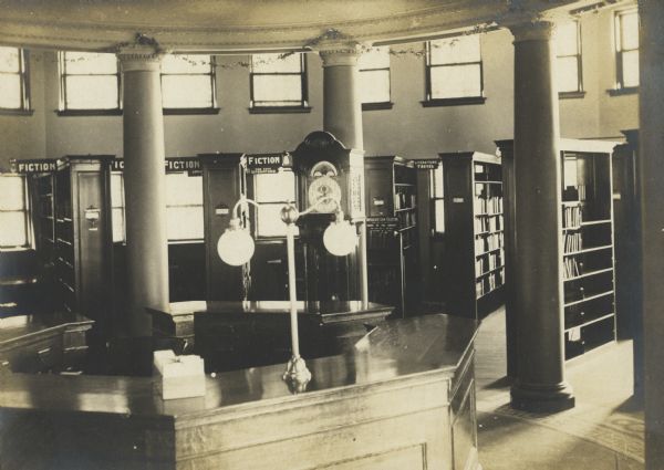 Interior view of the Stephenson Public Library. The library opened in 1903 funded with a gift from Isaac Stephenson of $30,000. Reverse of cardboard backing reads: "The stack room - from the charging desk - Stephenson Public Library, Marinette Wis." The room has a circular layout, and in the foreground is a large octagon librarian's desk with card file, surrounded by columns. Beyond the desk are bookshelves with the labels: Fiction, Language, Fine Arts Literature, Literature Travel, and Duplicate Loan Collection.