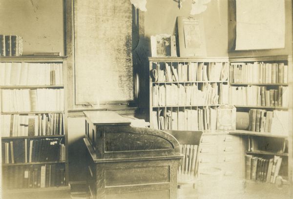 Interior view of the Stephenson Public Library. Reverse on the cardboard backing reads: "Built 1903, cost $30,000, Patton, Miller, Chicago, arch." In the foreground is a librarian's desk, a cabinet with drawers, and bookshelves along the wall. A window with its shade drawn is behind the desk.