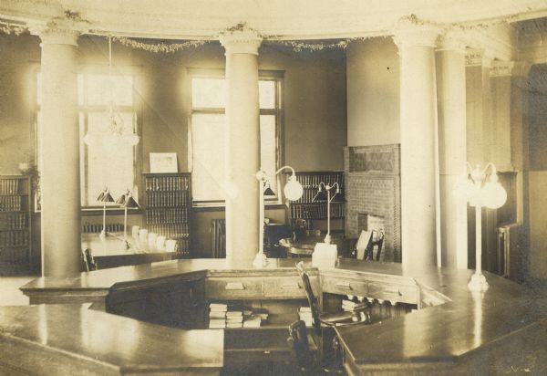 Interior view of the Stephenson Public Library. In the foreground is a large octagon librarian's desk surrounded by columns. Bookshelves are along the wall. There are also reading tables with lamps, and a fireplace on the right.