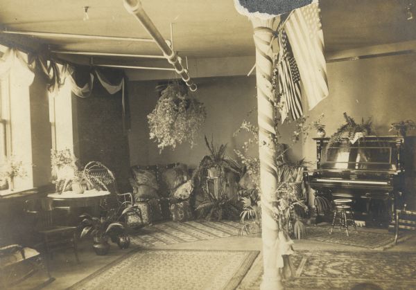 Interior view of the Stephenson Public Library. Cardboard backing reads: "Marinette - Lecture room - Stephenson Public Library." Reverse of cardboard backing reads: "Lecture room decorated for Woman's Club reception to teachers. Oct 23, 1903." In the foreground is a decorated column, with flags attached. On the wall on the right is an upright piano and stool. Potted plants and ferns are on the floor, tables, and hanging from the ceiling. A couch with pillows, and a rocking chair are in the left corner.