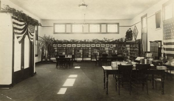Interior view of the Medford Public Library. Reverse of photograph reads: "Medford Wis, Feb 1917." On the left a flag, and a pine branch swag, adorn the double-door entrance. In the room are reading tables, bookshelves against the walls, a large potted fern, and another flag on the wall on the right. More pine branches are arrayed on top of the bookshelves just above a row of prints.