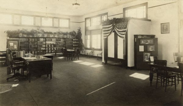 Interior view of the Medford Public Library. Library funded with a $6,000 gift from Andrew Carnegie. On the right a flag, and a pine branch swag, adorn the double-door entrance. In the room are reading tables, a desk, and bookshelves against the walls. More pine branches are arrayed on top of the bookshelves just above a row of prints.