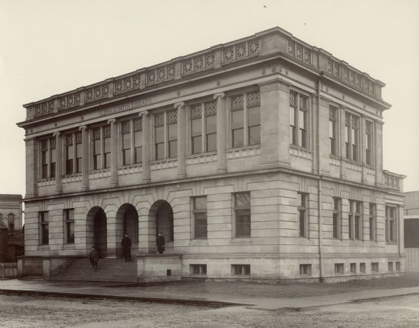 Exterior view from road of the Elisha D. Smith Library. The library opened in 1898 funded with a gift of $20,000 from E.D. Smith. Over the main entrance, at the roof line, it reads: "Elisha D. Smith Library." The building is made of stone and has three large stone arches at the entrance. Three men are standing on the front steps.