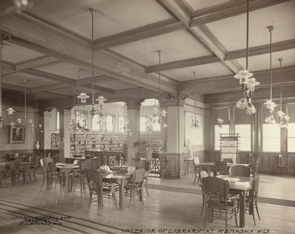 Interior view of the Elisha D. Smith Library. The library opened in 1898 funded with a gift of $20,000 from E.D. Smith. Lower left of photograph reads: "Van Ryn & de Gelleke Architects, Milwaukee, Wis." and lower right reads: "Interior of the Library at Menasha Wis." Throughout the room in the foreground are reading tables and chairs. Another room beyond columns and a wood carved railing are bookshelves and a card catalog. Other items in the room are a statue of Venus De Milo, a newspaper rack, and lamps suspended from the exposed beam ceiling.