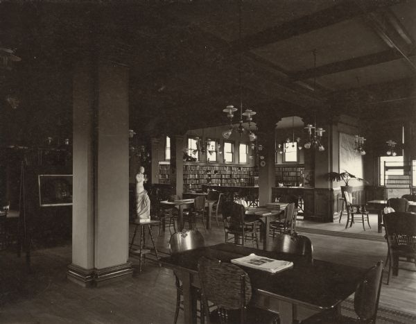 Interior view of the Elisha D. Smith Library. The library opened in 1898 funded with a gift of $20,000 from E.D. Smith. Throughout the room in the foreground are reading tables and chairs, a librarian's desk and card catalog. Another room beyond columns and a wood carved railing are bookshelves and a card catalog. Other items in the room are a statue of Venus De Milo, a newspaper rack, and lamps suspended from the exposed beam ceiling.