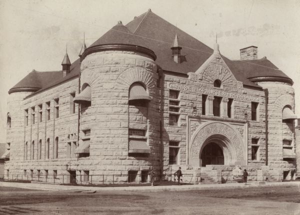 Exterior view of the Mabel Tainter Memorial Library. The library opened in 1891 and was funded by a $95,000 gift from Captain & Mrs. A. Tainter. Detailed carved stone archway over the main entrance that reads: "1889." Above the columns it reads: "Mabel Tainter Memorial." Richardsonian Romanesque style stone building with turrets. Three boys stand in front of the library near a railing, and striped awnings are over the windows.<p>Additional notes from "Some Wisconsin Library Buildings, 1904." Population 5,655. Borrowers 2,547. Circulation 31,978. Volumes 8,536. Tax $400 This building, housing the Memorial Free Library, was erected in 1889, at a cost, fully equipped, of $125,000, a gift to the public from Capt. and Mrs. Andrew Tainter, of Menomonie, as a memorial of their daughter.</p>