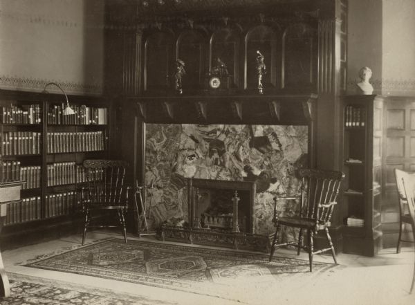 Interior view of the Mabel Tainter Memorial Library. The library opened in 1891 and was funded by a $95,000 gift from Captain & Mrs. A. Tainter. Dominating the room is a marble fireplace with an elaborately carved wood mantel. Rugs, chairs, and bookshelves surround the fireplace.