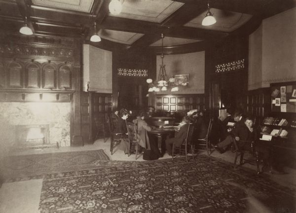Interior view of the Mabel Tainter Memorial Library. The library opened in 1891 and was funded by a $95,000 gift from Captain & Mrs. A. Tainter. On the right is a marble fireplace with an elaborately carved wood mantel, and area rugs cover the floor. A group of people are sitting at a reading table, men sit in chairs on the right near the wall, and a young boy sits near a book rack in the right foreground. Lamps hang from an exposed beam ceiling.