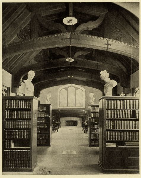Interior view of the Greene Memorial Library of Milwaukee-Downer College (now the University of Wisconsin-Milwaukee). The image is part of a 14-page pamphlet created by Hammersmith Engraving Co., Milwaukee, to advertise Downer College. View is down aisle through bookshelves, with a fireplace on the far wall. Reading tables and chairs are also at the back of the room. Busts are displayed on top of the bookshelves. The room has a high, arched ceiling with exposed ceiling beams.