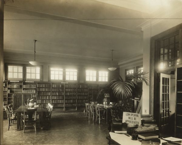 Interior view of the South Milwaukee Public Library. Sign on librarian's desk reads: "Low speaking will be appreciated." Signs on bookshelves read: "Natural Science 500. Useful Arts 600, and Fiction." In the room are reading tables and chairs, a librarian's desk with boxes of cards, and a large potted plant. Lamps hang from the tall ceiling, and a row of windows is along the top of the bookshelves.