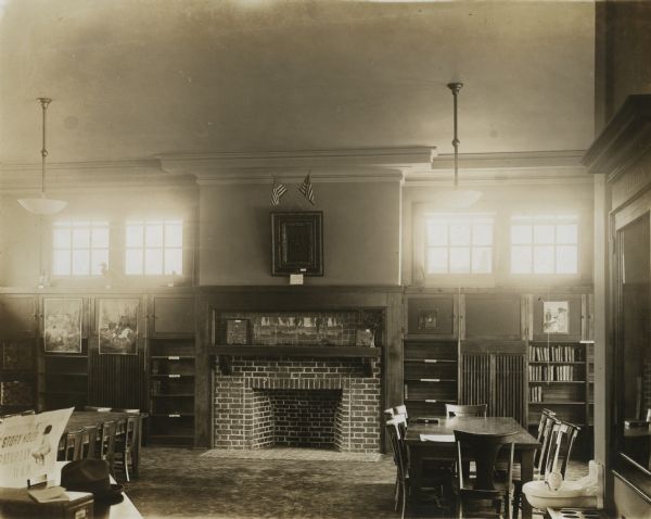 Interior view of the South Milwaukee Public Library. A sign on a table reads: "Story Hour Saturday 11 A.M." There is a large, brick fireplace along the far wall, which is flanked by windows above the bookshelves. There is a water fountain on the right, and reading tables and chairs are in the foreground.