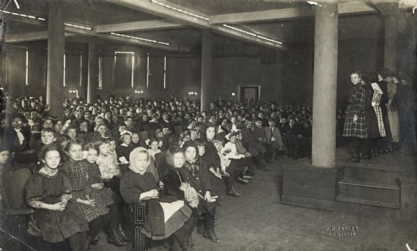 Interior view of the South Milwaukee Public Library. Reverse of cardboard backing reads: "South Milwaukee branch: a children entertainment." Stamped into photograph: "J.H. Taylor Milwaukee." A group of children is on stage on the right facing a large audience, which is made up mostly of children, with some adults scattered throughout. Most of the audience is looking at the camera.
