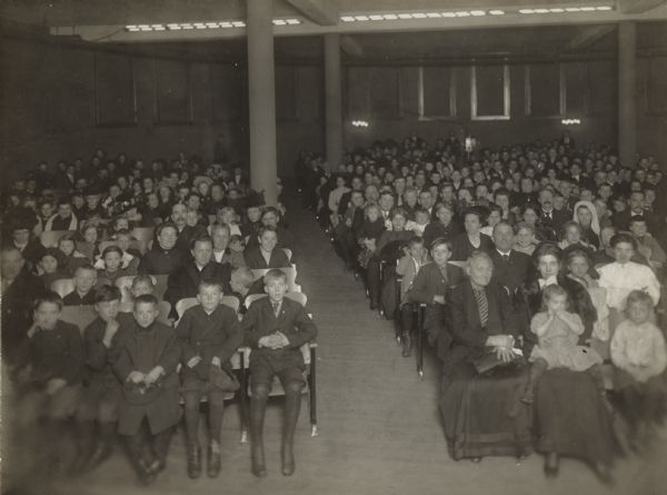 Interior view of the South Milwaukee Public Library of an audience of children and adults from the stage area of an auditorium.
