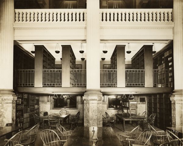 Interior view of the Milwaukee Public Library, showing reading tables on the ground floor, and balconies and railings of two levels above. Large stone columns with marble bases reach up the three floors.