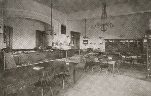Interior view of the Milwaukee Public Library. Note below the image reads: " Milwaukee Public Library, Children's Room." There are reading tables and chairs in the foreground. A long counter is along the back wall in an arched niche. Bookshelves are along the right wall, and lamps and chandeliers hang from the high ceiling.