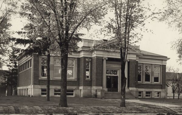 Exterior view of the Arabut Ludlow Memorial Library. The library opened in 1905 and was funded with a $13,930 donation from H.E. & W. Ludlow. This image is part of a seven-image hand-bound booklet. On the interior of the front flap it reads: "The Arabut Ludlow Memorial Library, Monroe, Wisconsin, Claude K. Stark, Architects, Madison, Wis, Photographs by E.H. Gloege, Monroe Wis." The image is stamped: "E.H. Gloege Monroe, Wis." Above the main entrance of the brick and stone building it reads: "Free Library." There is fresh sod on the lawn in front.