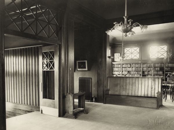 Interior view of the Arabut Ludlow Memorial Library. The library opened in 1905 and was funded with a $13,930 donation from H.E. & W. Ludlow. This image is part of a seven-image hand-bound booklet. On the interior of the front flap it reads: "The Arabut Ludlow Memorial Library, Monroe, Wisconsin, Claude K. Stark, Architects, Madison, Wis, Photographs by E.H. Gloege, Monroe Wis." The image is stamped: "E.H. Gloege Monroe, Wis." Near the entrance is a large, wood bench, and a chandelier. Bookshelves, reading tables and chairs can be seen in the room behind.