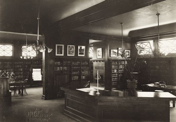 Interior view of the Arabut Ludlow Memorial Library. The library opened in 1905 and was funded with a $13,930 donation from H.E. & W. Ludlow. This image is part of a seven-image hand-bound booklet. On the interior of the front flap it reads: "The Arabut Ludlow Memorial Library, Monroe, Wisconsin, Claude K. Stark, Architects, Madison, Wis, Photographs by E.H. Gloege, Monroe Wis." The image is stamped: "E.H. Gloege Monroe, Wis." In the foreground is a three-sided librarian's desk, and a chandelier. Bookshelves line the walls. Large, open doorways open into another room, in which can be seen a fireplace, a newspaper rack, reading tables and chairs.