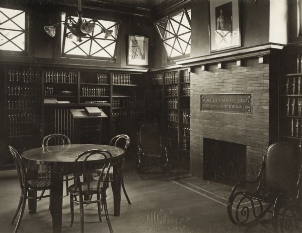 Interior view of the Arabut Ludlow Memorial Library. The library opened in 1905 and was funded with a $13,930 donation from H.E. & W. Ludlow. This image is part of a seven-image hand-bound booklet. On the interior of the front flap it reads: "The Arabut Ludlow Memorial Library, Monroe, Wisconsin, Claude K. Stark, Architects, Madison, Wis, Photographs by E.H. Gloege, Monroe Wis." The image is stamped: "E.H. Gloege Monroe, Wis." There is a brick fireplace at the right of the photograph with a plaque over it that reads: "Arabut Memorial Library / erected in loving memory by his family 1904." Bookshelves line the walls of the room, a rocking chair is to the right of the fireplace, and there is a round reading table with chairs.