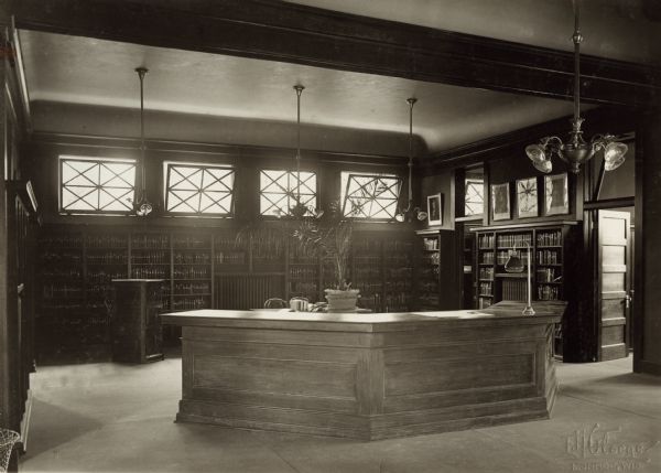 Interior view of the Arabut Ludlow Memorial Library in Monroe, WI. The library opened in 1905 and was funded with a $13,930 donation from H.E. & W. Ludlow. This image is part of a seven-image hand-bound booklet. On the interior of the front flap it reads: "The Arabut Ludlow Memorial Library, Monroe, Wisconsin, Claude K. Stark, Architects, Madison, Wis, Photographs by E.H. Gloege, Monroe Wis." The image is stamped: "E.H. Gloege Monroe, Wis." In the foreground is a three-sided librarian's desk. Behind it bookshelves line the walls, and windows are in a row above them along the back wall.