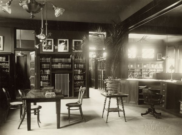 Interior view from behind the librarian's desk of the Arabut Ludlow Memorial Library. The library opened in 1905 and was funded with a $13,930 donation from H.E. & W. Ludlow. This image is part of a seven-image hand-bound booklet. On the interior of the front flap it reads: "The Arabut Ludlow Memorial Library, Monroe, Wisconsin, Claude K. Stark, Architects, Madison, Wis, Photographs by E.H. Gloege, Monroe Wis." The image is stamped: "E.H. Gloege Monroe, Wis." There is a reading table with chairs, and a large potted plant on a table in the foreground. On the back wall are bookshelves and a door to another room. Beyond the librarian's desk on the right is another room with bookshelves along the walls.
