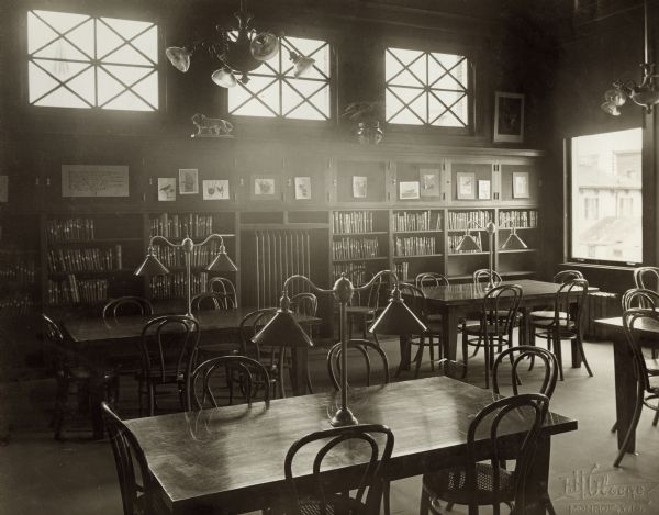Interior view of the Arabut Ludlow Memorial Library. The library opened in 1905 and was funded with a $13,930 donation from H.E. & W. Ludlow. This image is part of a seven-image hand-bound booklet. On the interior of the front flap it reads: "The Arabut Ludlow Memorial Library, Monroe, Wisconsin, Claude K. Stark, Architects, Madison, Wis, Photographs by E.H. Gloege, Monroe Wis." The image is stamped: "E.H. Gloege Monroe, Wis." In the foreground are reading tables with lamps, bookshelves along the back wall. Buildings are visible through a large window on the right.