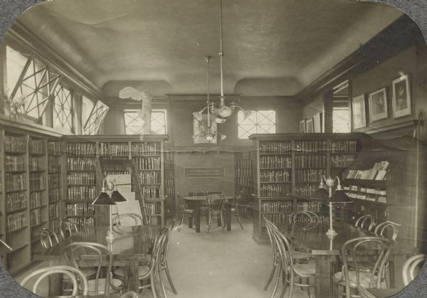 Interior view of the Arabut Ludlow Memorial Library. The library opened in 1905 and was funded with a $13,930 donation from H.E. & W. Ludlow. There is a brick fireplace with a plaque over it that reads: "Arabut Memorial Library, erected in loving memory of his family 1904." In the room are reading tables and chairs, bookshelves, a statue of Winged Victory, a newspaper rack, and chandeliers and reading lamps.