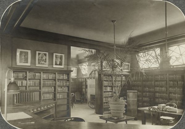 Interior view of the Arabut Ludlow Memorial Library. The library opened in 1905 and was funded with a $13,930 donation from H.E. & W. Ludlow. In the foreground is the top of a three-sided librarian's desk. Along the walls are bookshelves and a doorway opens into another room with a fireplace.