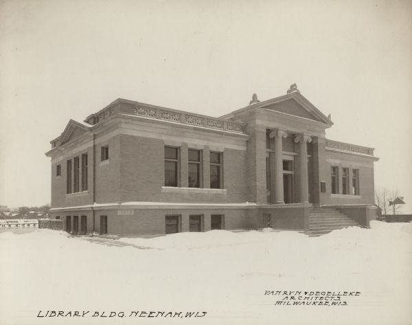 Exterior view of the Neenah Public Library in winter. The library opened in 1904 and was funded with a combined donation of $20,000 from the citizens of Neenah and Andrew Carnegie. Above the main entrance it reads: "Public Library." Cornerstone displays the year 1902. Notes in ink on the photograph. Lower left reads: "Library Bldg. Neenah, Wis," and lower right reads: "Van Ryn & DeGelleke Architects, Milwaukee, Wis." The building is of brick and stone, and has ionic columns, and a pediment at the entrance.