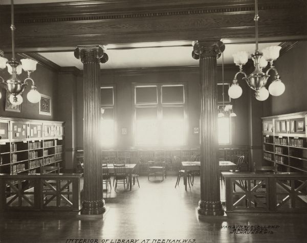 Interior view of the Neenah Public Library. The library opened in 1904 and was funded with a combined donation of $20,000 from the citizens of Neenah and Andrew Carnegie. Notes in ink on the photograph. Lower left reads: "Interior of the Library at Neenah, Wis," and lower right reads: "Van Ryn & DeGelleke Architects, Milwaukee, Wis." The library has hardwood floors and carved wood trim. There is a decorative wood railing with two ionic columns framing a room with reading tables and bookshelves and windows along the back wall.