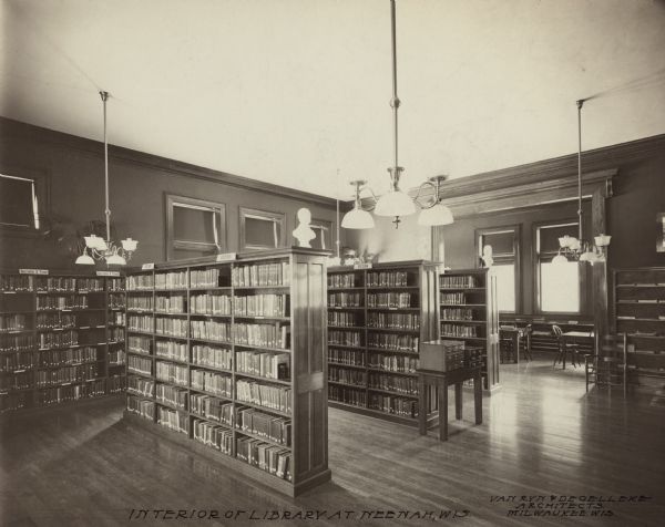 Interior view of the Neenah Public Library. The library opened in 1904 and was funded with a combined donation of $20,000 from the citizens of Neenah and Andrew Carnegie. Notes in ink on the photograph. Lower left reads: "Interior of the Library at Neenah, Wis," and lower right reads: "Van Ryn & DeGelleke Architects, Milwaukee, Wis." The library has hardwood floors, and chandeliers hang from the tall ceiling. There are freestanding bookshelves as well as bookshelves along the walls. Some have signs for "Fiction" and "Description & Travel." There are two small card catalogs, and a second room is visible with reading tables and chairs.