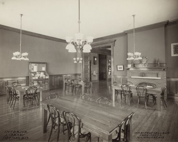 Interior view of the Neenah Public Library. The library opened in 1904 and was funded with a combined donation of $20,000 from the citizens of Neenah and Andrew Carnegie. Notes in ink on the photograph. Lower left reads: "Interior Library, Neenah, Wis," and lower right reads: "Van Ryn & DeGelleke Architects, Milwaukee, Wis." In the large, open room are wood reading tables and bentwood chairs, and a magazine and display rack. There are chandeliers hanging from the tall ceiling. A large doorway opens into another room or hallway with a sink on the wall and a wall clock.