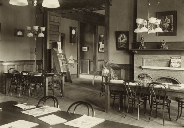 Interior view of the Neenah Public Library. The library opened in 1904 and was funded with a combined donation of $20,000 from the citizens of Neenah and Andrew Carnegie. Reverse of cardboard backing reads: "Reading Room." In the room are wood reading tables with bentwood chairs, a fireplace, a plant on a stand, and chandeliers hanging from the ceiling. Through a large doorway is a wall sink, and a wall clock.