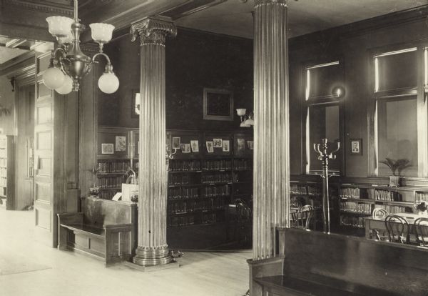 Interior view of the Neenah Public Library. The library opened in 1904 and was funded with a combined donation of $20,000 from the citizens of Neenah and Andrew Carnegie. Ink note on photograph reads: "Children's room." In the foreground is a chandelier, and two large benches on each side of two wood ionic columns. The room behind has bookshelves, a coat rack, and children's reading tables. On the back wall are windows with the shades drawn.